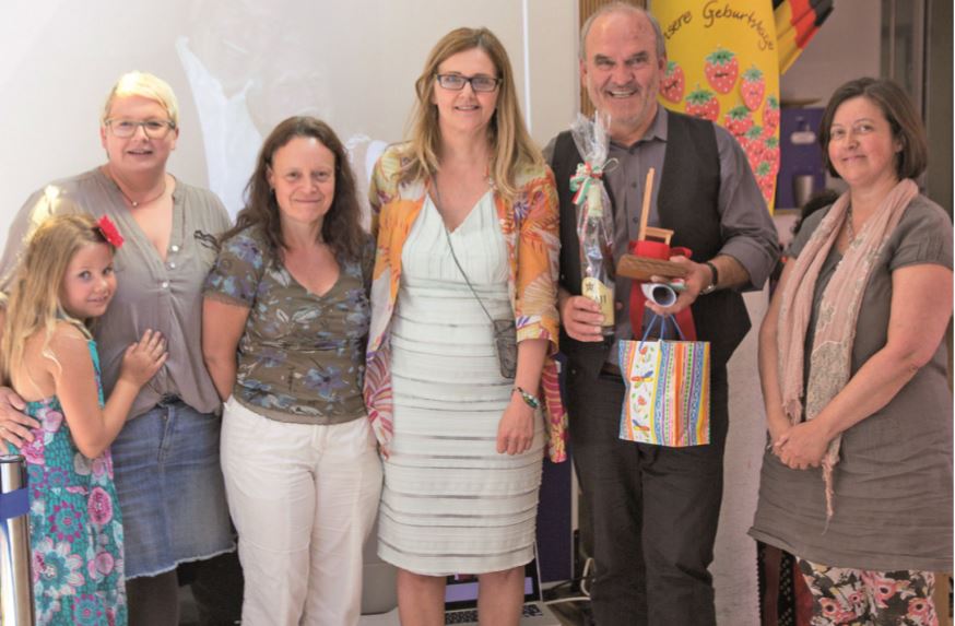 Mehrere Personen lächeln in die Kamera, ein Mann hält einen kleinen Stuhl und eine Flasche in Geschenkverpackung