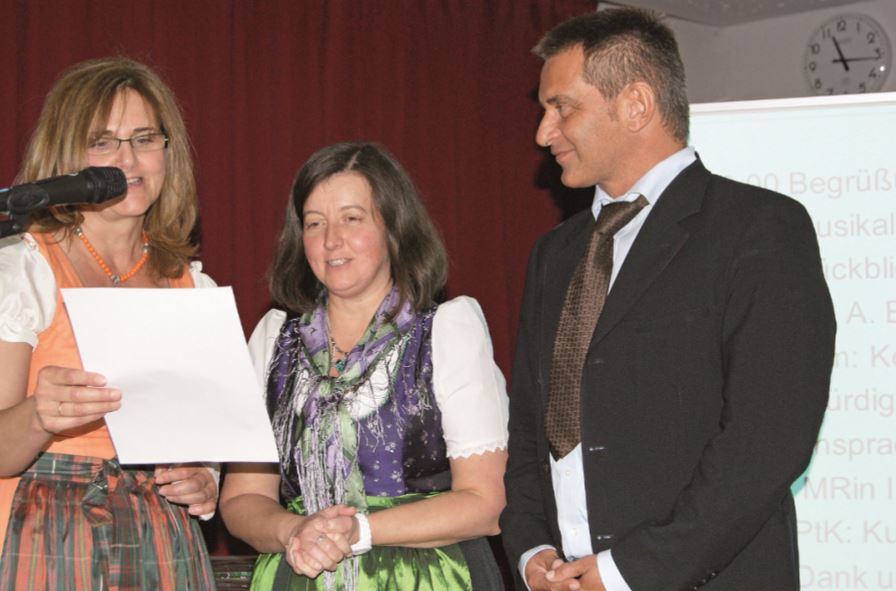 Zwei Frauen im Dirndl sprechen in Mikro eine hält ein weißes Blatt Papier und ein Mann im Anzug beobachtet sie