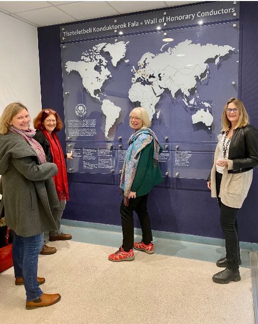 Vier Frauen stehen lächelnd vor einer Weltkarte mit Überschrift Tiszteletbeli Konduktorok Fala Wall of Honorary Conductors