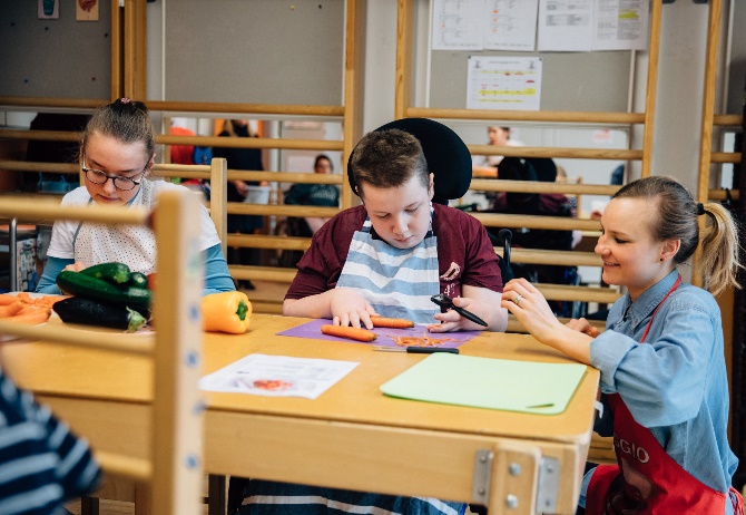 Zwei junge Personen schneiden Gemüse und ältere Frau kniet daneben und beobachtet sie