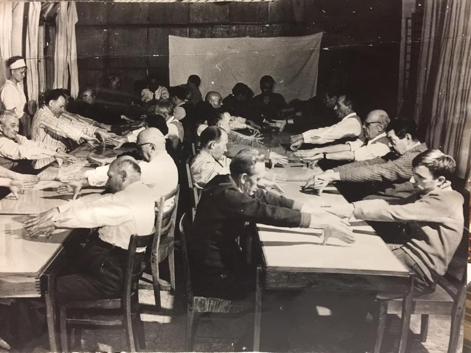 Schwarz-weiß: Viele Männer sitzen an langen Tischen gegenüber und strecken ihre Arme so aus dass der Daumen die Tischplatte berührt