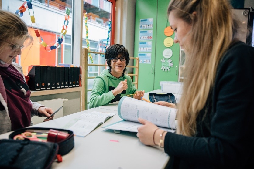 Zwei junge Personen sitzen an einem Tisch und eine ältere Frau blättert in einem Heft