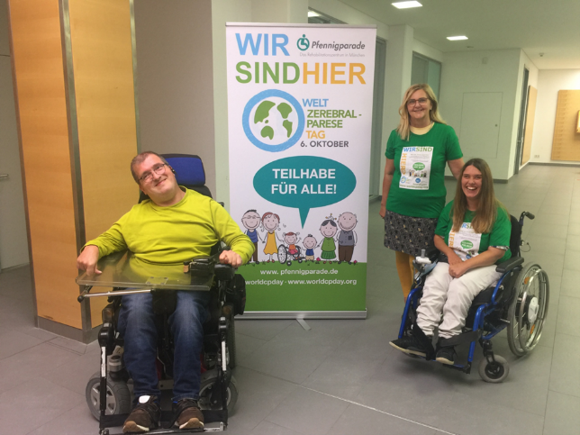 Aufsteller Wir sind hier Logo Pfennigparade Welt Zerebralparese Tag 6. Oktober Teilhabe für alle davor zwei Frauen in grünen Shirts und ein Mann in hellgrünem Pullover