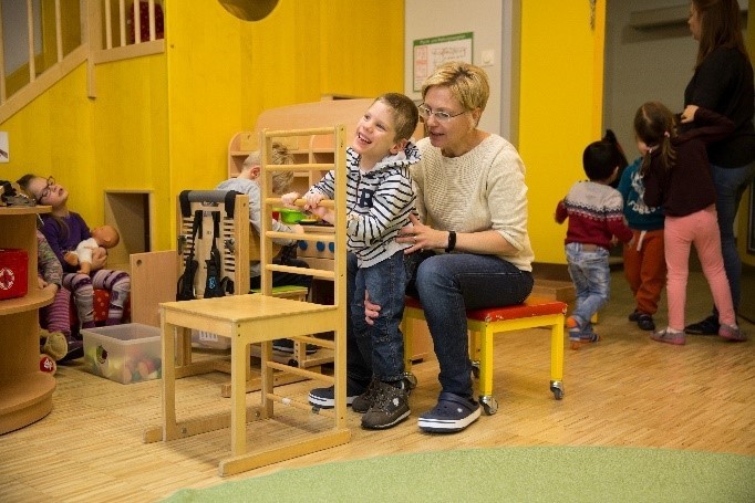 Foto: das Kind hält sich an der Stuhllehne fest und der Therapeut unterstützt es.