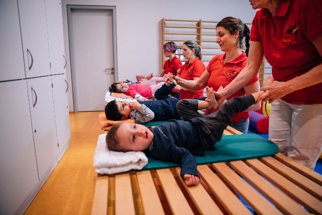 Foto: Eine große Holzbank, auf der kleine Kinder liegen und Therapeuten Übungen zum Festhalten der Kinderbeine durchführen.