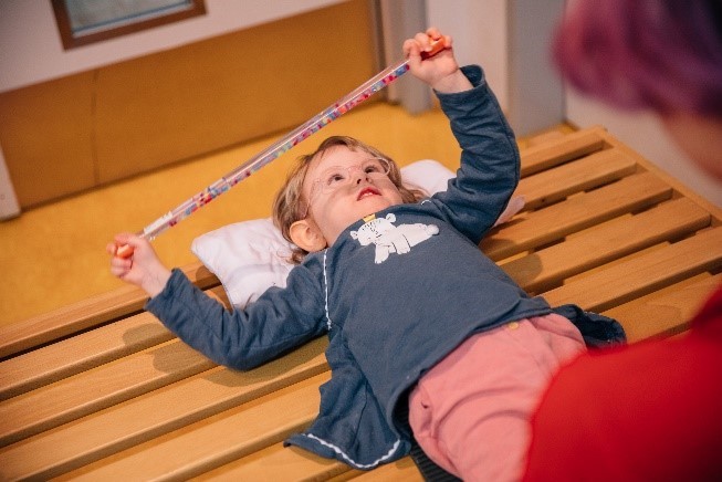 Foto: Ein kleines Mädchen liegt auf einer Holzbank und macht eine Übung. Sie hält einen bunten Stab in ihren Händen.